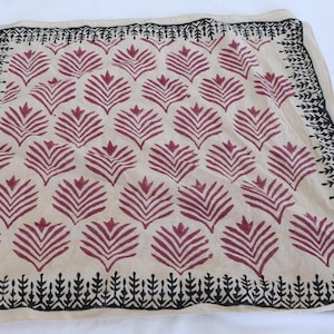 May include: A square, beige textile with a repeating pattern of maroon leaf-like shapes. The border is black with a repeating leaf design. The textile is likely a scarf or a decorative cloth.