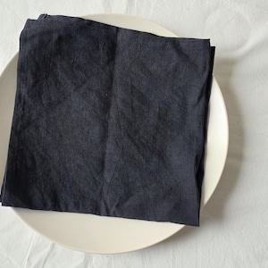 Peut inclure: Une serviette en lin bleu marine pliée repose sur une assiette en céramique blanche. L'assiette est centrée sur une nappe blanche texturée. La texture de la serviette contraste avec l'assiette lisse.