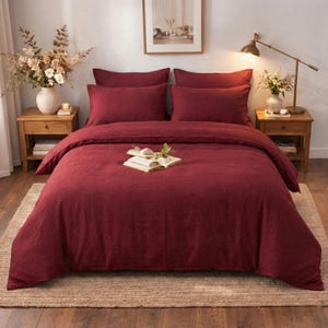 Puede incluir: Una cama con una funda nórdica de color rojo intenso y fundas de almohada a juego. La ropa de cama está colocada sobre una cama con dos mesitas de noche de madera a ambos lados. Un libro con vegetación descansa sobre la funda nórdica.