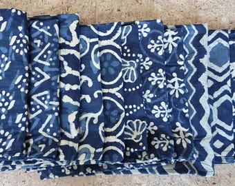 Puede incluir: Una pila de tela azul índigo doblada con varios diseños blancos. Los patrones incluyen formas florales, geométricas y abstractas. La tela parece ser de algodón o una fibra natural similar.