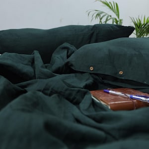 Può includere: Un set di biancheria da letto in lino verde scuro con chiusura a bottone sulle federe. La biancheria da letto è disposta su un letto con un diario in pelle marrone e una penna blu in cima.