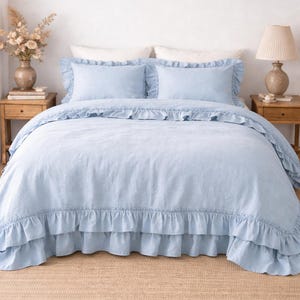 Peut inclure: Un ensemble de lit bleu clair avec des bords à volants, comprenant une housse de couette et deux taies d'oreiller. La literie est sur un lit avec un tapis beige et des tables de chevet en bois de chaque côté.