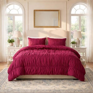 May include: A bed with a burgundy comforter and matching pillow shams. The comforter has a ruched design. The bed is in a bedroom with white nightstands, lamps, and arched windows. A gold framed mirror hangs above the bed.