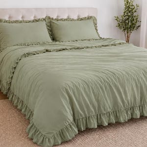 May include: Sage green bed set with frilled edges, including a duvet cover and two pillowcases. The bedding is on a bed with a beige headboard. A lamp and side table are visible in the background.