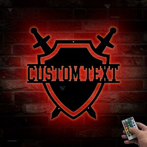 May include: A black metal shield with two swords crossed behind it. The shield has the text "CUSTOMTEXT" written across the center. The shield is illuminated with red light. A remote control is visible in the bottom right corner.