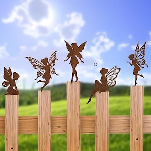 May include: Five brown metal fairy silhouettes are mounted on a wooden fence. Each fairy has detailed wings and is posed differently, with one kneeling, one dancing, and another seated. The background features a green field and a blue sky with clouds.