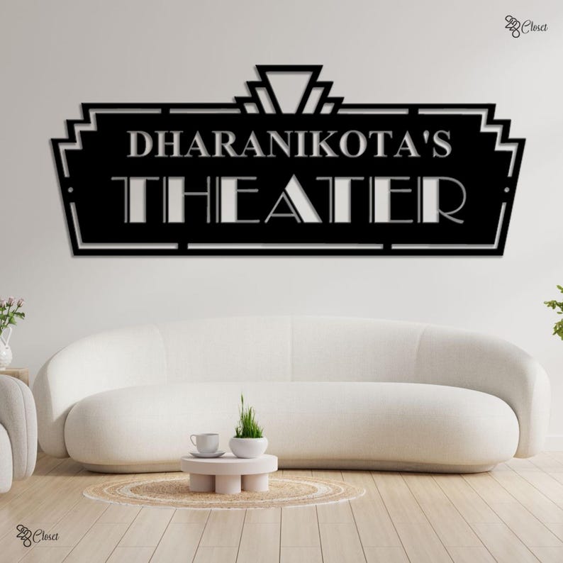 May include: Black metal wall art sign that reads "DHARANIKOTA'S THEATER" in a stylized font. The sign has an art deco design and is mounted on a white wall above a white sofa. The sign is a decorative piece.