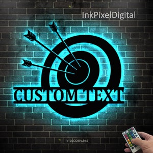 May include: Black metal target with three arrows and the text "CUSTOM TEXT" in a split monogram style. The target is illuminated with a blue light and mounted on a brick wall. A remote control with multiple colored lights is visible in the bottom right corner.