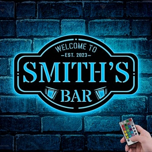 May include: Black metal sign with a blue neon glow, reading "Welcome to Smith's - Est. 2023 - Bar". The sign is mounted on a brick wall. A remote control with multiple colored buttons is visible in the bottom right corner.