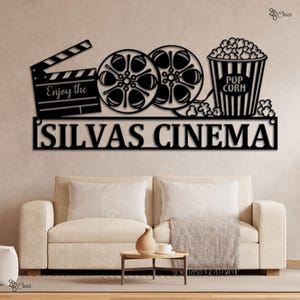 May include: Black metal wall art featuring a movie clapperboard, film reels, and a popcorn bucket. The text "Enjoy the" is on the clapperboard, and "SILVAS CINEMA" is below. The art is mounted above a beige sofa.