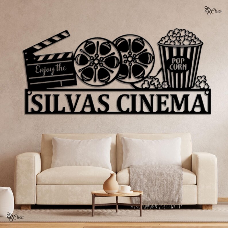 May include: Black metal wall art featuring a movie clapperboard, film reels, and a popcorn bucket. The text "Enjoy the" is on the clapperboard, and "SILVAS CINEMA" is below. The art is mounted above a beige sofa.
