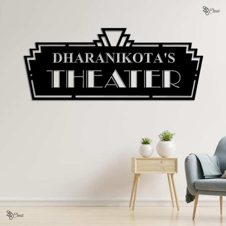 May include: Black metal wall art with the words "DHARANIKOTA'S THEATER" in a decorative font. The sign has an art deco design and is mounted on a white wall. The image includes a small side table with plants.
