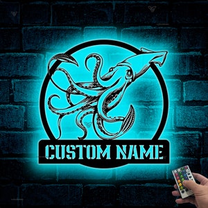 Op de afbeelding: Een zwarte metalen uitsnijding van een inktvis met tentakels in een cirkel met de woorden "CUSTOM NAME" eronder. De uitsnijding is verlicht met blauw licht en is gemonteerd op een bakstenen muur. Een afstandsbediening bevindt zich in de rechterbenedenhoek van de afbeelding.
