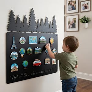 Puede incluir: Tablero magnético de metal negro con una silueta de bosque en la parte superior, que contiene imanes con temática de viajes. Los imanes representan monumentos, mapas y un globo terráqueo. Un niño alcanza un imán de globo aerostático. La cita "Not all those who wander are lost..." está en la parte inferior.