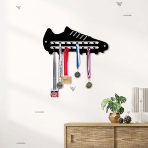 Puede incluir: Portamedallas de metal negro en forma de zapato con múltiples ganchos para colgar medallas. El soporte está montado en una pared blanca con un estante de madera debajo. Hay varias medallas colgadas de los ganchos, incluida una cinta roja, blanca y azul.