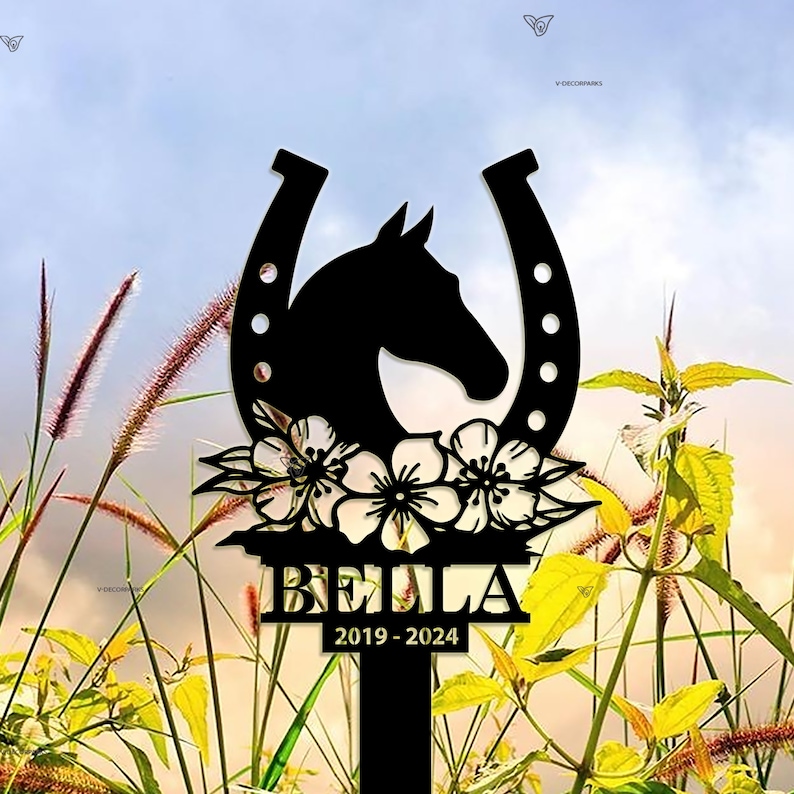 May include: Black metal silhouette of a horse's head inside a horseshoe with flowers and the name "BELLA" and the dates "2019-2024" below. The silhouette is in front of a blurred background of green grass and a blue sky.