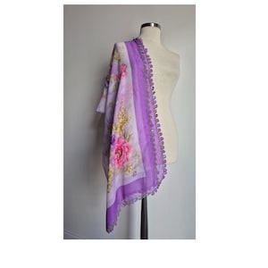 May include: A lightweight lavender scarf featuring a floral print with pink and yellow flowers. The scarf has a solid purple border and is finished with a delicate, dark purple crocheted lace trim along the edge, draped over a mannequin.