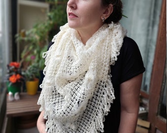 Pode incluir: Um xale triangular de crochê na cor creme com um padrão rendado intrincado, detalhes texturizados em relevo e uma borda decorativa com franjas. O xale está drapeado ao redor do pescoço e ombros, exibindo o trabalho detalhado do fio.