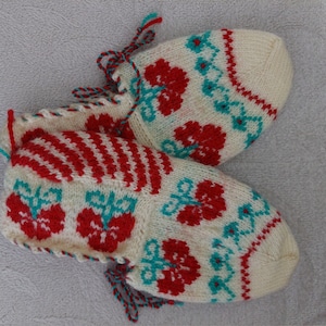 Puede incluir: Par de zapatillas blancas de punto con motivos florales rojos y azul verdoso. Las zapatillas tienen un borlón decorativo.