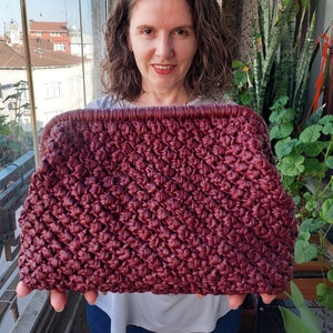 May include: A close-up of a burgundy crocheted bag held by a person. The bag is made with a textured stitch and has a simple, rectangular shape.