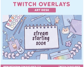 May include: A blue and white digital art illustration of an art desk with a notepad that says "stream starting soon". The desk is decorated with paintbrushes, paint tubes, a watercolor palette, a boba tea keychain, a flower charm, and a small dog charm.
