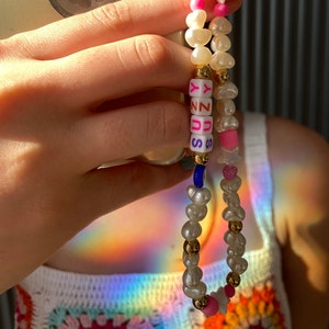 May include: A beaded phone charm with white, pink, and gold details. The charm includes irregular pearls, star-shaped beads, and letter beads spelling "SUNNY". A blue bead and pink heart-shaped beads are also included.