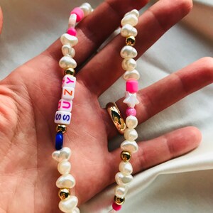 May include: A handmade necklace with freshwater pearls, gold-coloured beads, and colourful accents. The necklace spells "SUNNY" with letter beads, and includes pink and blue beads and a star charm. The necklace is held in a hand.