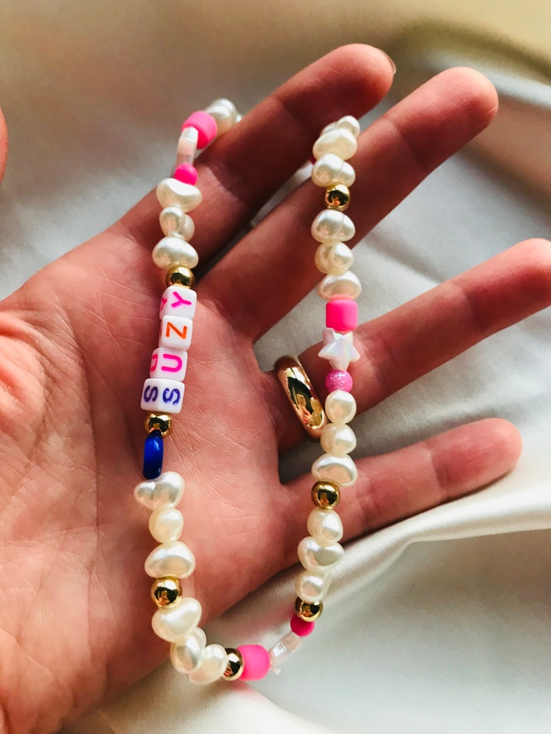 May include: A handmade necklace with freshwater pearls, gold-coloured beads, and colourful accents. The necklace spells "SUNNY" with letter beads, and includes pink and blue beads and a star charm. The necklace is held in a hand.