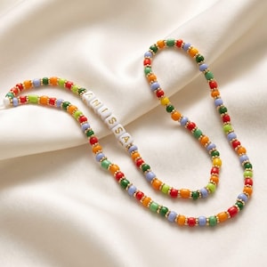May include: A colorful beaded necklace with the name "ANISSA" spelled out in white letter beads. The necklace features a variety of small, round beads in red, orange, yellow, green, and blue, separated by gold-colored spacers. The necklace is displayed on a white satin surface.