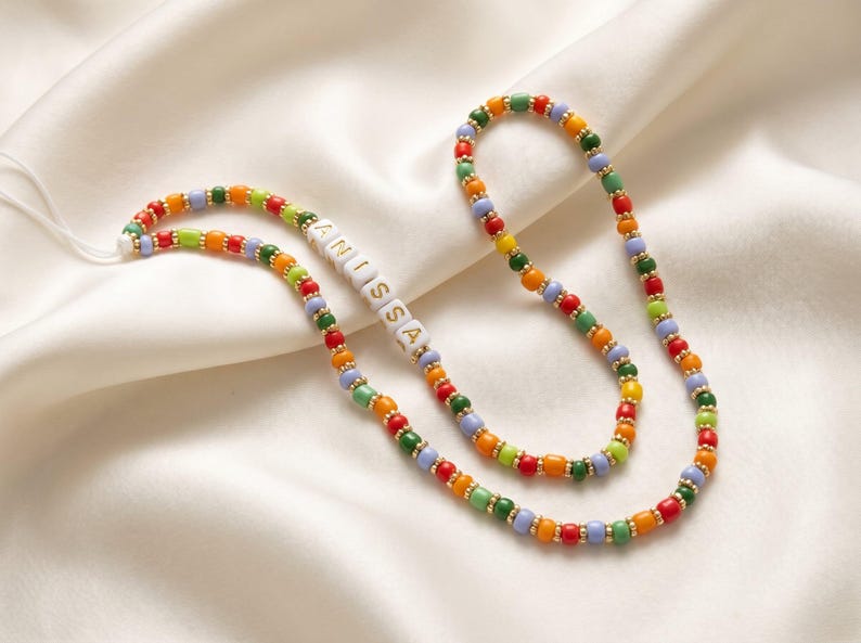 May include: A colorful beaded necklace with the name "ANISSA" spelled out in white letter beads. The necklace features a variety of small, round beads in red, orange, yellow, green, and blue, separated by gold-colored spacers. The necklace is displayed on a white satin surface.