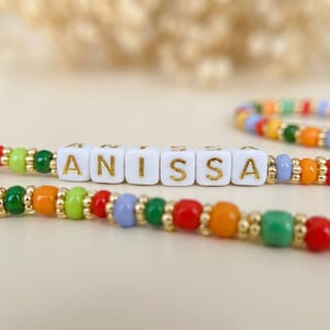 May include: A colorful beaded bracelet with the name "ANISSA" spelled out in gold letters on white square beads. The bracelet features red, orange, green, and blue beads. A second bracelet with similar colors is in the background.