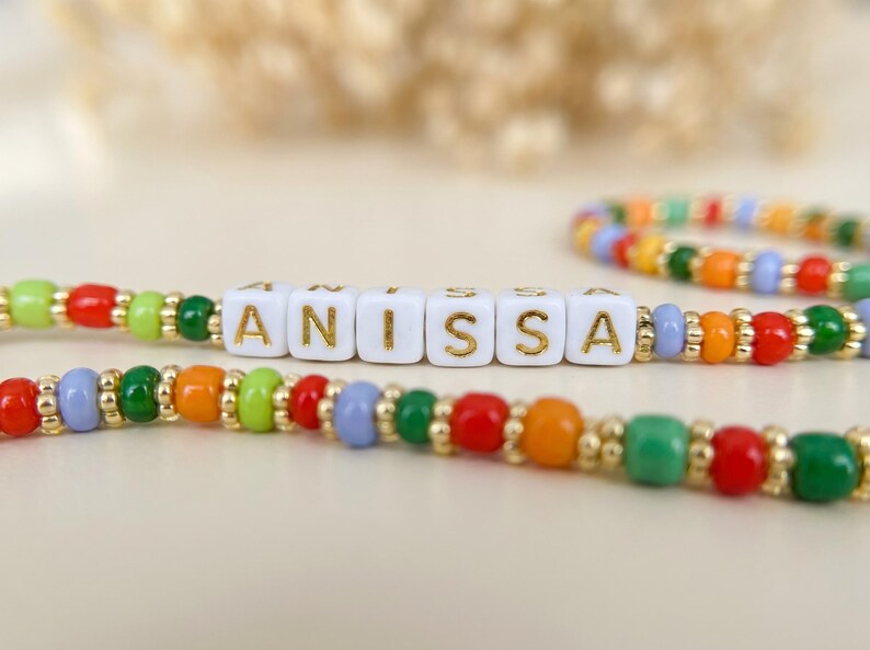 May include: A colorful beaded bracelet with the name "ANISSA" spelled out in gold letters on white square beads. The bracelet features red, orange, green, and blue beads. A second bracelet with similar colors is in the background.