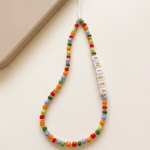 May include: A colorful beaded phone charm with the name "ANISSA" spelled out in white letter beads. The charm features a variety of small beads in red, orange, yellow, green, and blue, with gold-colored spacer beads. A white cord connects the charm to a phone.