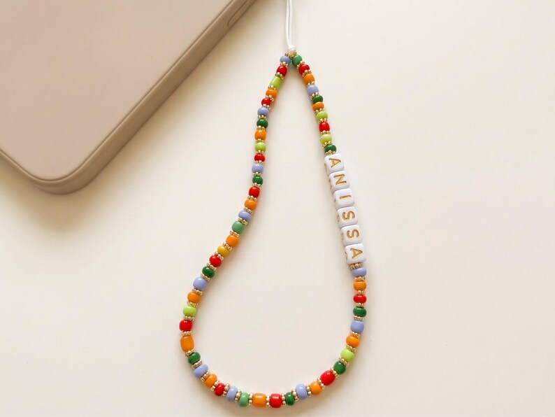 May include: A colorful beaded phone charm with the name "ANISSA" spelled out in white letter beads. The charm features a variety of small beads in red, orange, yellow, green, and blue, with gold-colored spacer beads. A white cord connects the charm to a phone.