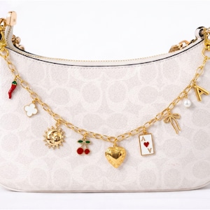 May include: A white crescent-shaped handbag with a subtle repeating logo pattern, featuring a gold-tone chain strap adorned with various charms including a red chili pepper, a sun, cherries, a heart, an ace of hearts card, a bow, and the letter A.