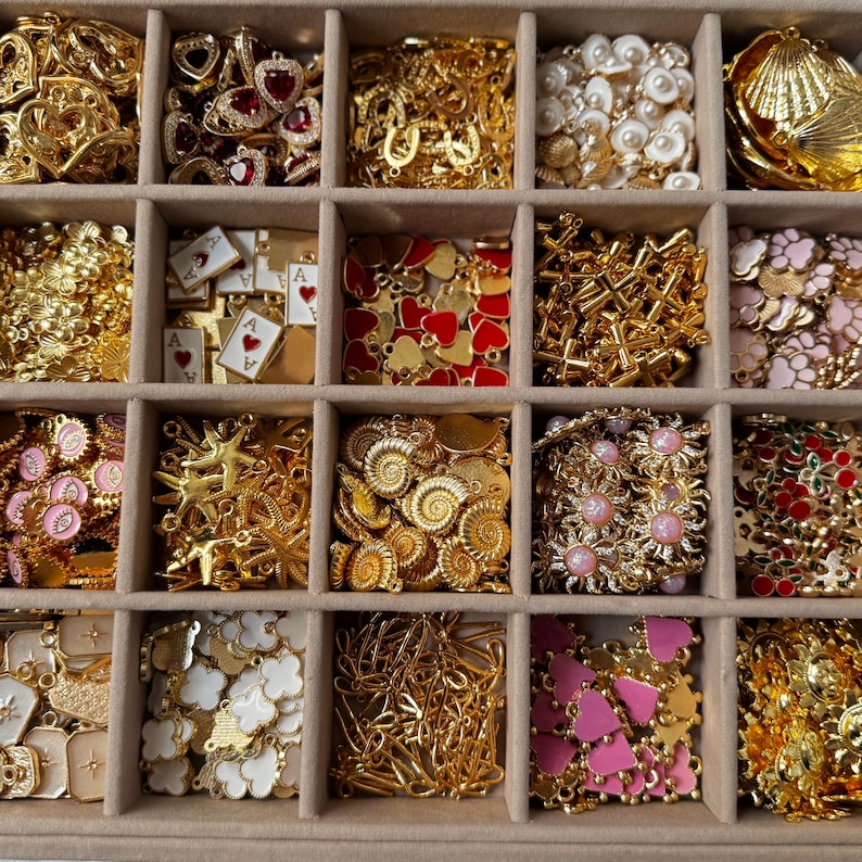 May include: A variety of gold-toned jewelry findings and charms, including hearts, shells, stars, and floral designs, are arranged in a divided tray. Some charms feature red and pink enamel accents, and playing card charms are also visible.