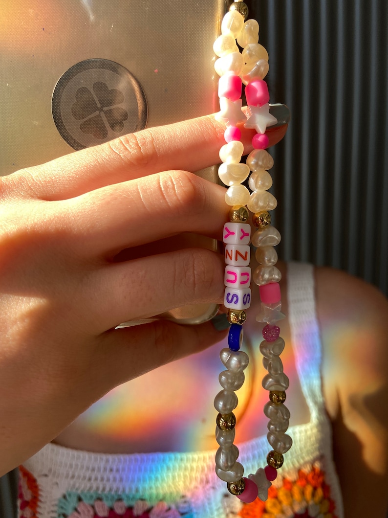 May include: A beaded phone charm with white, pink, and gold details. The charm includes irregular pearls, star-shaped beads, and letter beads spelling "SUNNY". A blue bead and pink heart-shaped beads are also included.