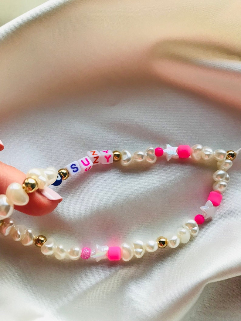 May include: A bracelet made of white pearl beads, gold accents, and bright pink and white star-shaped beads. The name "SUZY" is spelled out in colourful letter beads. The bracelet is a delicate piece of jewellery.