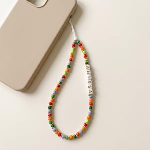 May include: A colorful beaded phone charm with the name "ANISSA" spelled out in white letter beads. The charm features a variety of beads in red, green, orange, and blue, with gold-colored accents. A beige phone case is in the background.