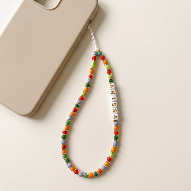 May include: A colorful beaded phone charm with the name "ANISSA" spelled out in white letter beads. The charm features a variety of beads in red, green, orange, and blue, with gold-colored accents. A beige phone case is in the background.
