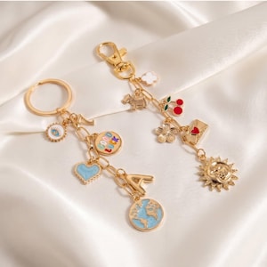 May include: Two gold-colored keychains with various charms, including a heart, globe, and sun. The charms are in various colors, including blue, red, and white. The keychains are on a white, silky background.