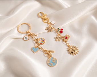 May include: Two gold-colored keychains with various charms, including a heart, globe, sun, and cherries. The charms are in shades of blue, red, and white. The keychains are on a white, silky background.