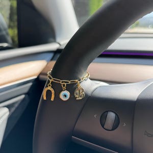 Op de afbeelding: Gouden bedelarmband met een hoefijzer, een boze oog en de letter "B" bedels. De armband wordt getoond op een zwart stuurwiel. De bedels zijn goudkleurig en de boze oog bedel heeft blauwe en witte details.