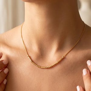 Puede incluir: Un collar de tono dorado que presenta una cadena delicada con un segmento central compuesto por pequeñas cuentas redondas y finas barras cilíndricas alternadas, creando un patrón lineal que descansa sobre la zona de la clavícula.