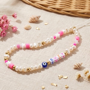 May include: A beaded phone charm featuring white pearls, gold beads, pink star and cylinder beads, and letter beads spelling "SUZY". An evil eye bead is also included. The charm is on a beige surface with flowers and seashells.