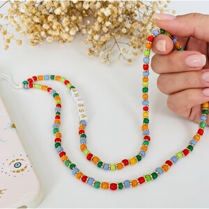 May include: A colorful beaded phone strap with red, orange, yellow, green, and blue beads. The strap has gold-colored spacers and the name "ANISSA" is printed on a white section. A pink phone case with eye designs is visible.