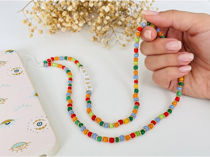 May include: A colorful beaded phone strap with red, orange, yellow, green, and blue beads. The strap has gold-colored spacers and the name "ANISSA" is printed on a white section. A pink phone case with eye designs is visible.