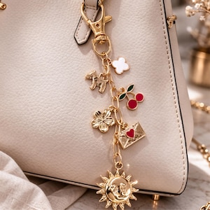 May include: A gold charm bracelet featuring a dog, a four-leaf clover, a cherry, a white flower, an envelope with a heart, and a sun with a face. The charms are suspended from a gold chain and clasp, attached to a cream-coloured handbag.