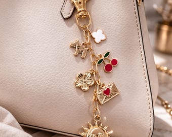 May include: A gold-colored charm bracelet with various charms, including a dog, a four-leaf clover, a cherry, a white flower, an envelope with a heart, and a sun with a face. The charms are attached to a gold chain and a clasp, hanging from a cream-colored handbag.