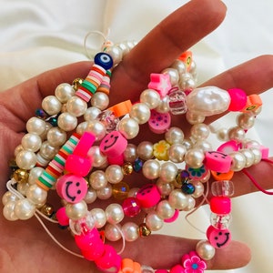 May include: Several beaded bracelets with white faux pearls, neon pink smiley faces, and colourful accent beads. Some bracelets include flower and star-shaped beads, and an evil eye charm.